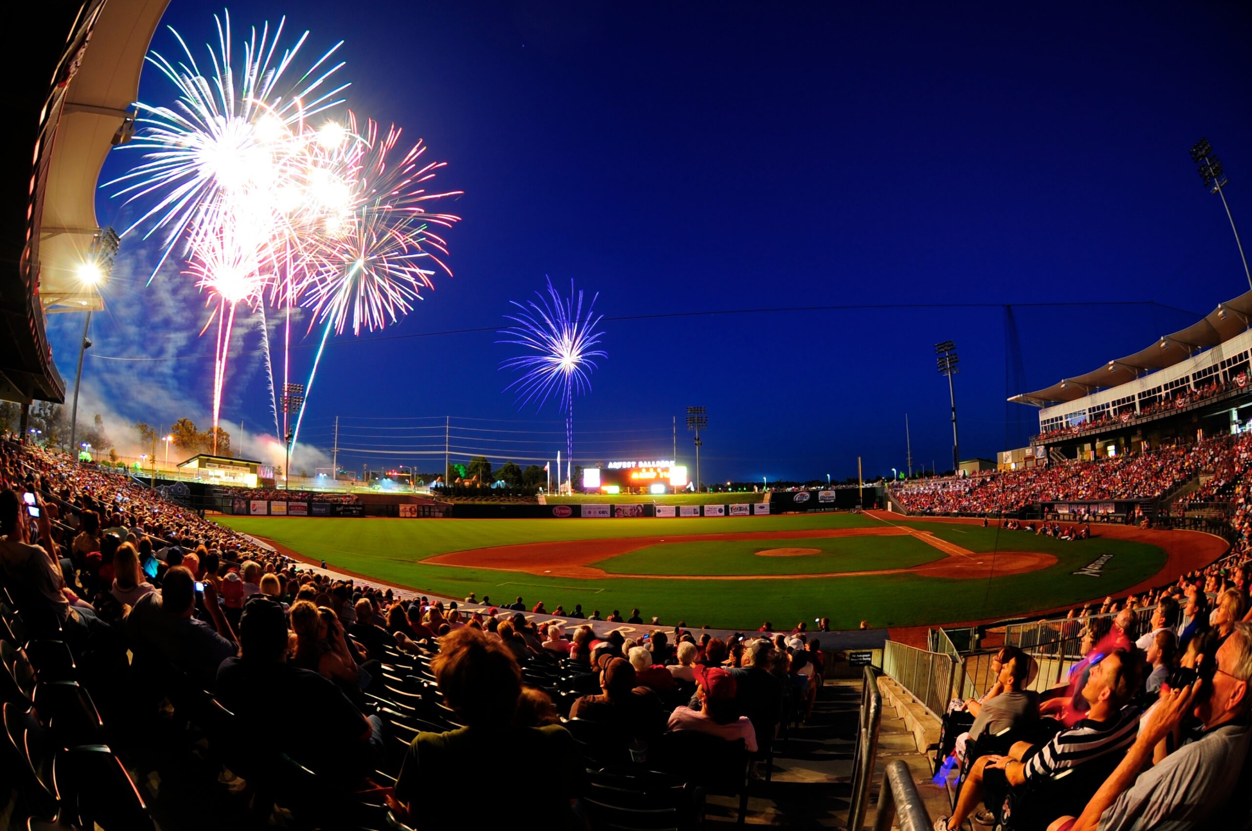 A large crowd at a baseball game sits in the bucket seats and watches fireworks shoot off at the far end of the baseball field.