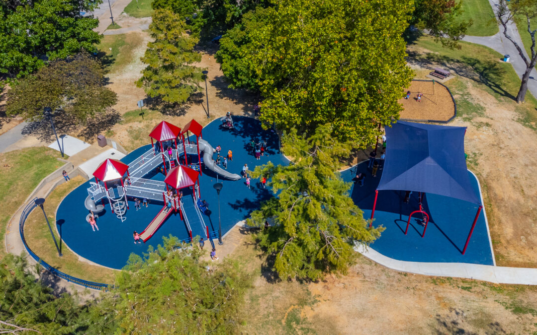 An aerial view of Murphy Park in Springdale. The park has a blue floor and canopy. The playground structure is red and white metal pipes with red roofs and gray slides.