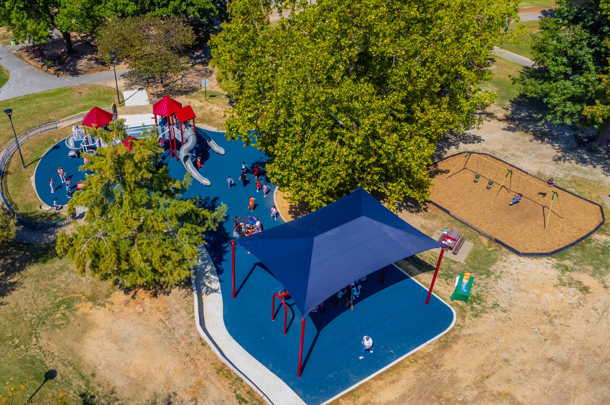 An aerial view of Murphy Park in Springdale. The park has a blue floor and canopy. The playground structure is red and white metal pipes with red roofs and gray slides.